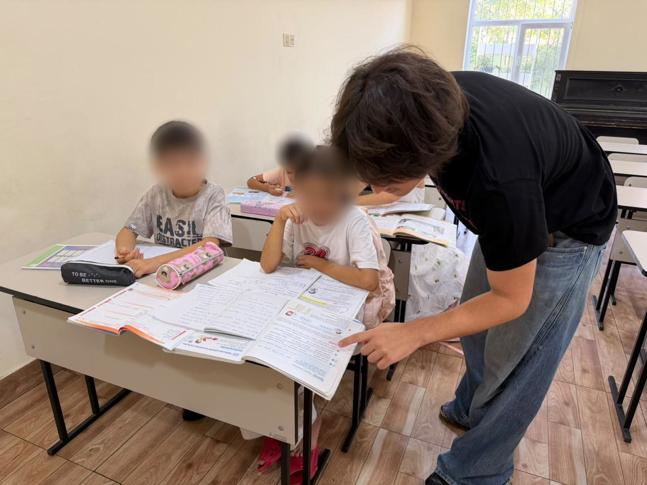 Jamal leans over a desk, pointing to a line in a girl's textbook while a boy looks on.